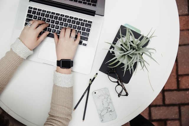 A person working on their blog on a laptop at a white table, beside a phone, glasses, pencils, and a small potted plant.