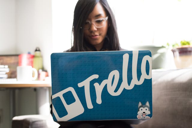 A woman using a blue laptop with the Trello logo visible on the back of the laptop.