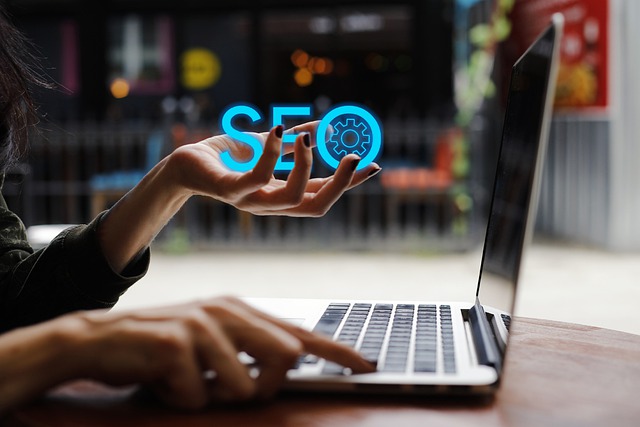 A person's hand holding the word "SEO" with a gear icon above a laptop keyboard, suggesting digital marketing and optimization.