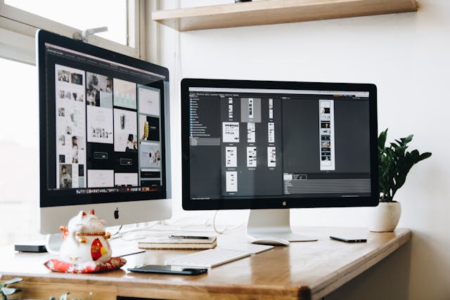 A minimalist workspace featuring two large desktop monitors displaying design software with various layouts and wireframes.