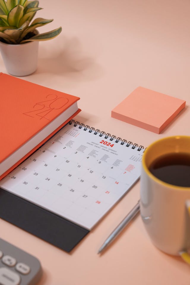 A 2024 spiral-bound calendar on a desk next to a notebook, a pen, a pink sticky note, a coffee mug, and a small potted plant.