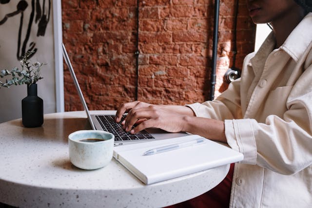 A person doing blog audit on their laptop at a table with a notebook, pen, coffee, and plant.