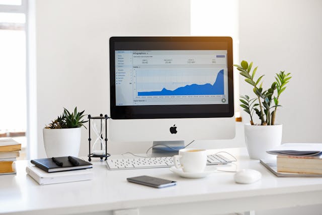 A workspace with an iMac displaying analytics charts, surrounded by plants, books, and an hourglass on a white desk.