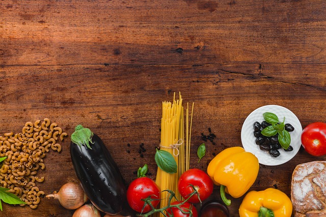 A wooden table with pasta, vegetables, and fresh ingredients, including eggplant, tomatoes, bell peppers, and basil.






