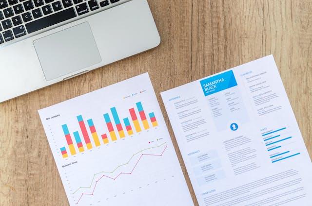 A laptop beside documents featuring a resume and colorful charts on a wooden desk.