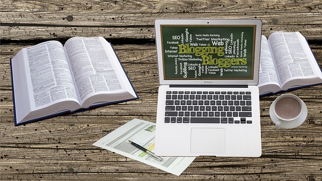 An open laptop displaying blogging-related keywords, surrounded by books, documents, and coffee.