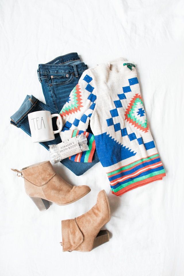 A flat lay of cozy fall fashion essentials: knit sweater, denim, ankle boots, and accessories.